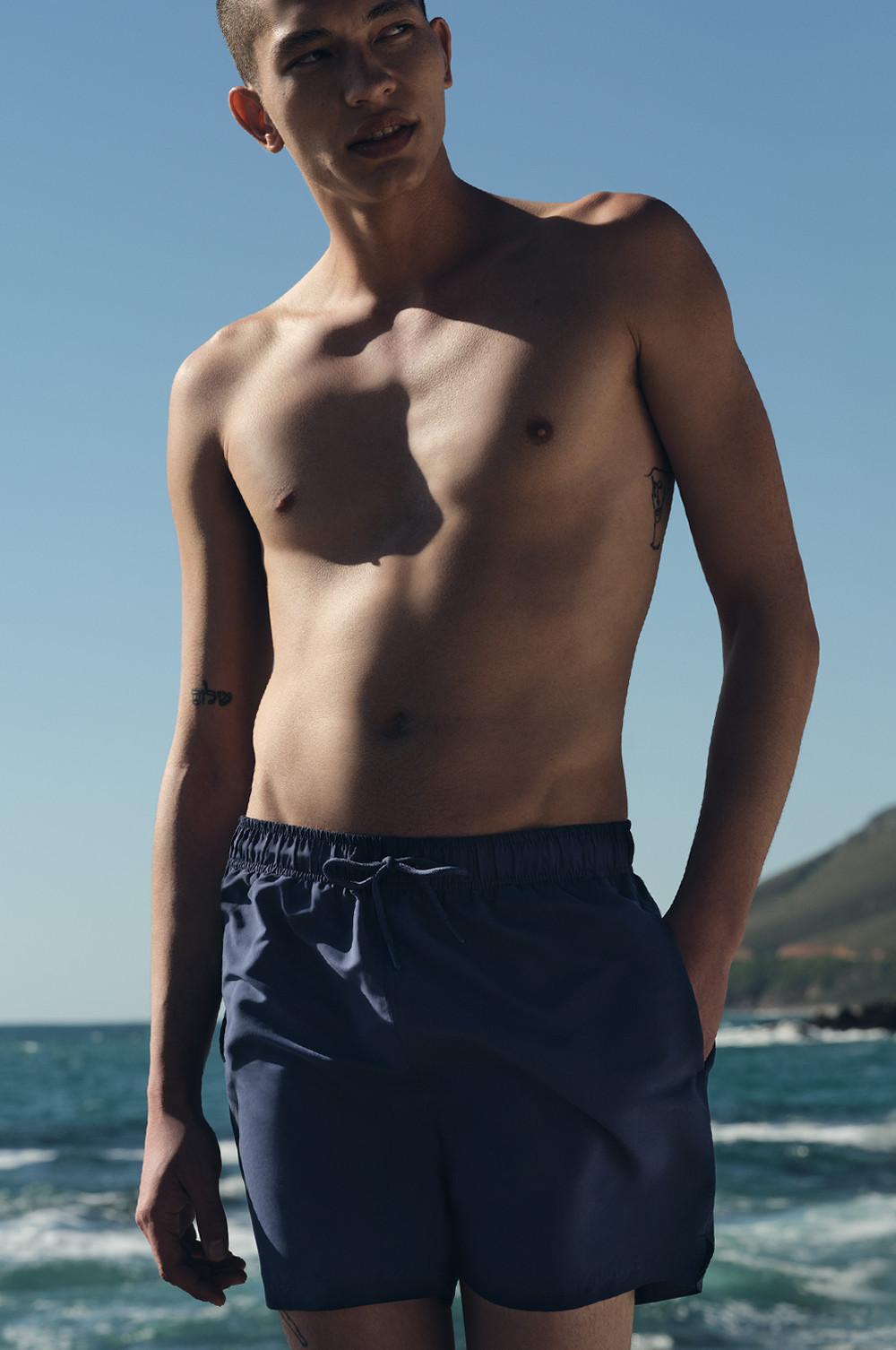 Un modello a torso nudo che indossa pantaloncini da bagno in microfibra blu navy, in posa con sullo sfondo l'oceano.