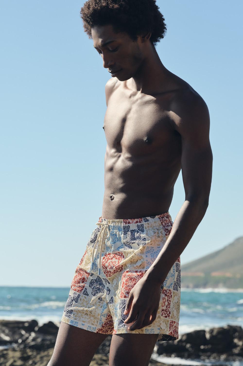 Pantaloncini da bagno a fantasia geometrica rossa, blu e panna, indossati da un modello su una spiaggia rocciosa con l'oceano sullo sfondo.