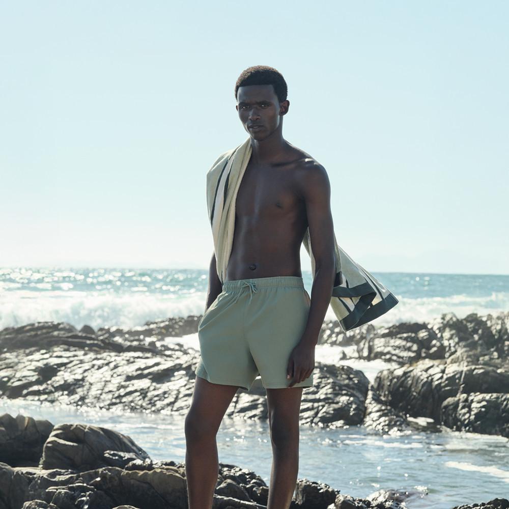 Modello che indossa pantaloncini da bagno verde menta e asciugamano beige sulle spalle, in piedi su una spiaggia rocciosa con le onde dell'oceano sullo sfondo.