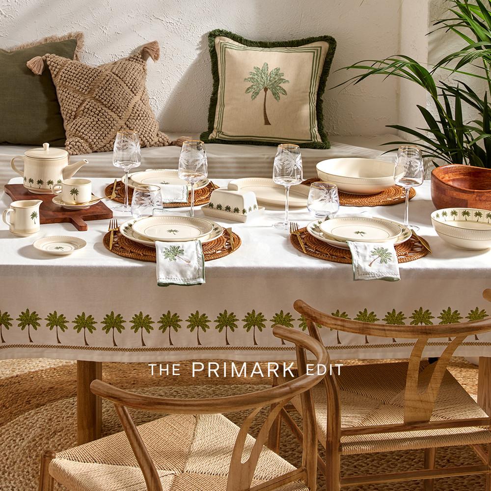 Elegante decoración de mesa con vajilla blanca, cojines a juego y mantel de Primark, todo con motivos de palmeras. Los salvamanteles tejidos completan el look.
