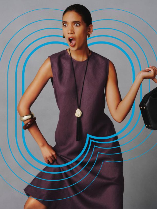 Person wearing mauve sports bra with black sweater raised above head, against pink background with white oval shape.