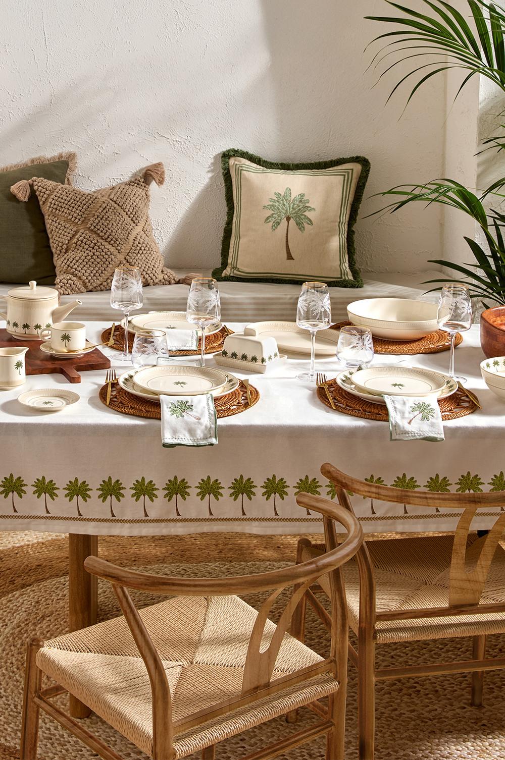 Decoración de mesa elegante con mantel, cojines y vajilla a juego con motivos de palmeras, manteles individuales de ratán y sillas de madera.