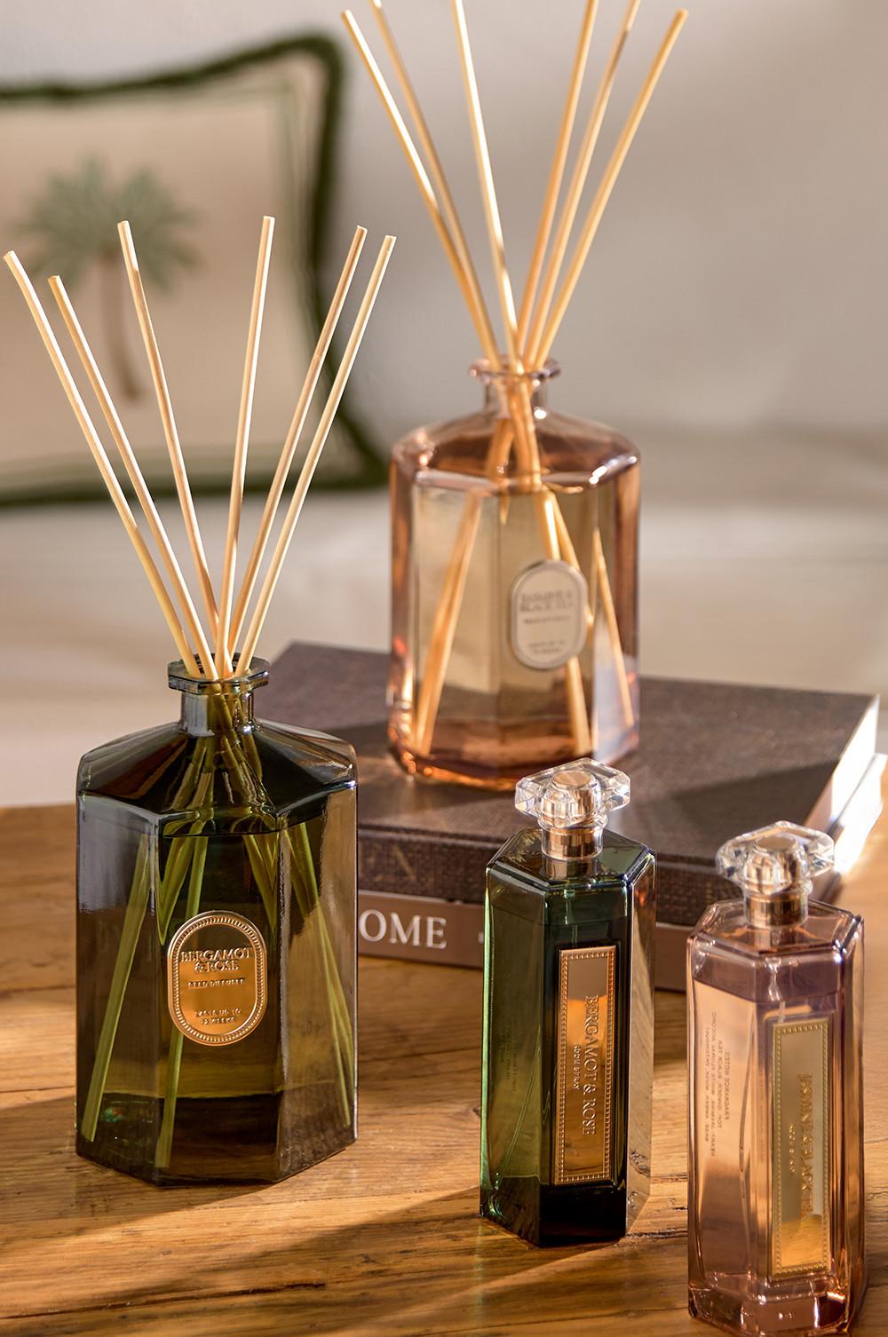 Colección de difusores de varillas y frascos de perfume de vidrio verde y ámbar con etiquetas doradas sobre una superficie de madera.