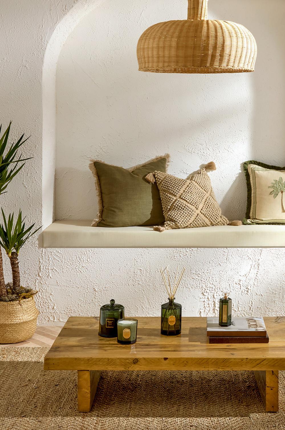 Banco de pared con cojines de macramé y de color verde oliva y mesa de centro de madera con velas bajo una lámpara colgante de mimbre.