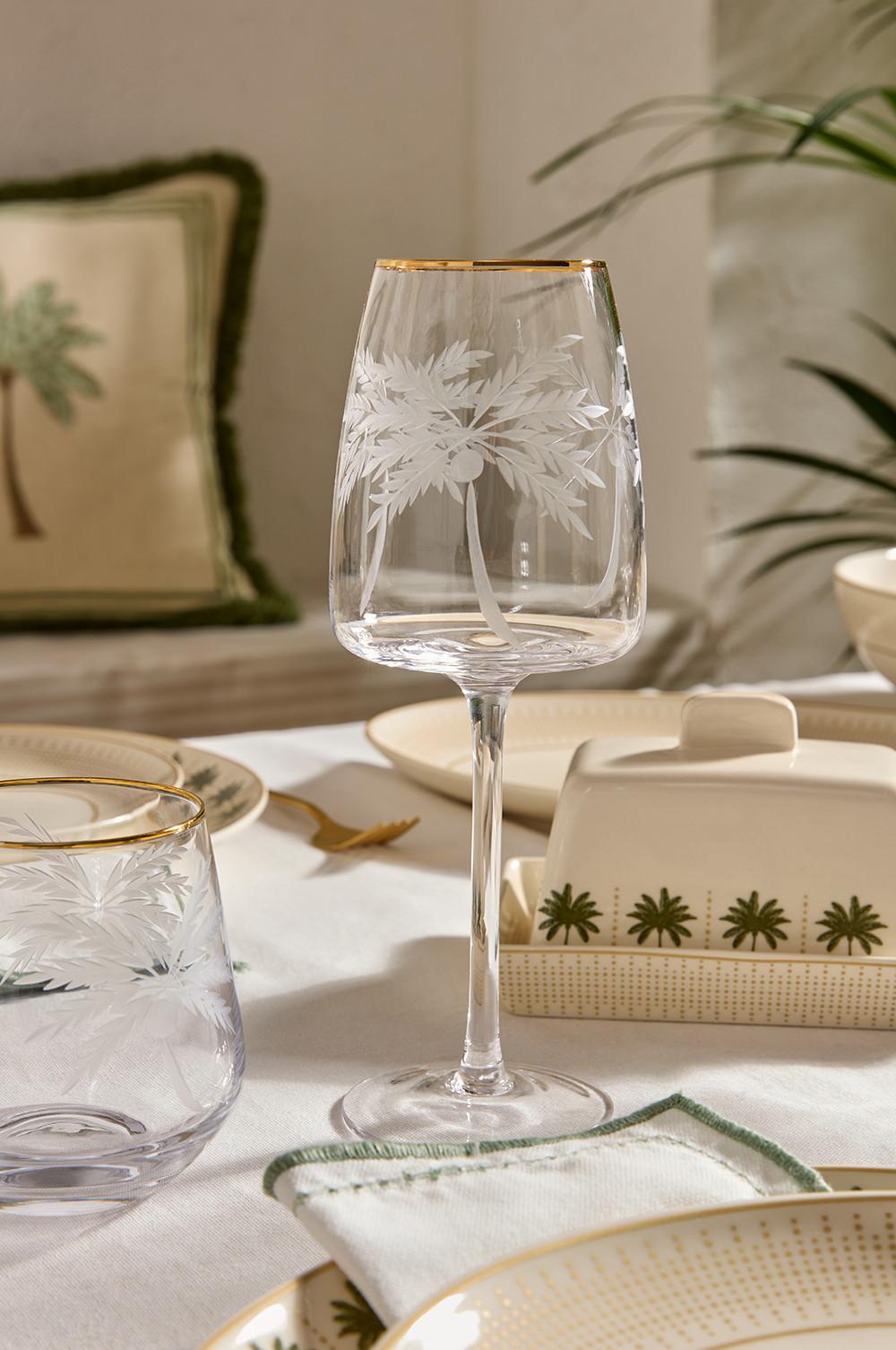 Copa de vino con diseño de palmera grabado y borde dorado sobre un mantel blanco con vajilla a juego.
