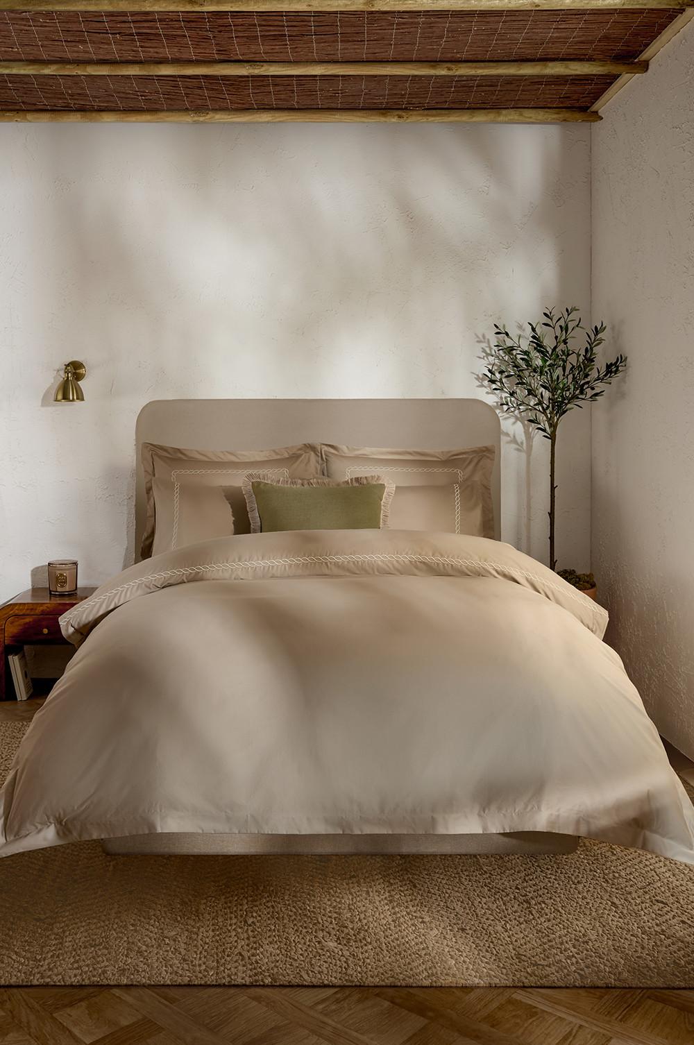 Ropa de cama blanca con camino de cama acolchado de color crema y cojines con bordados de palmeras y ribetes de color verde; junto a la cama hay una maceta con un olivo.