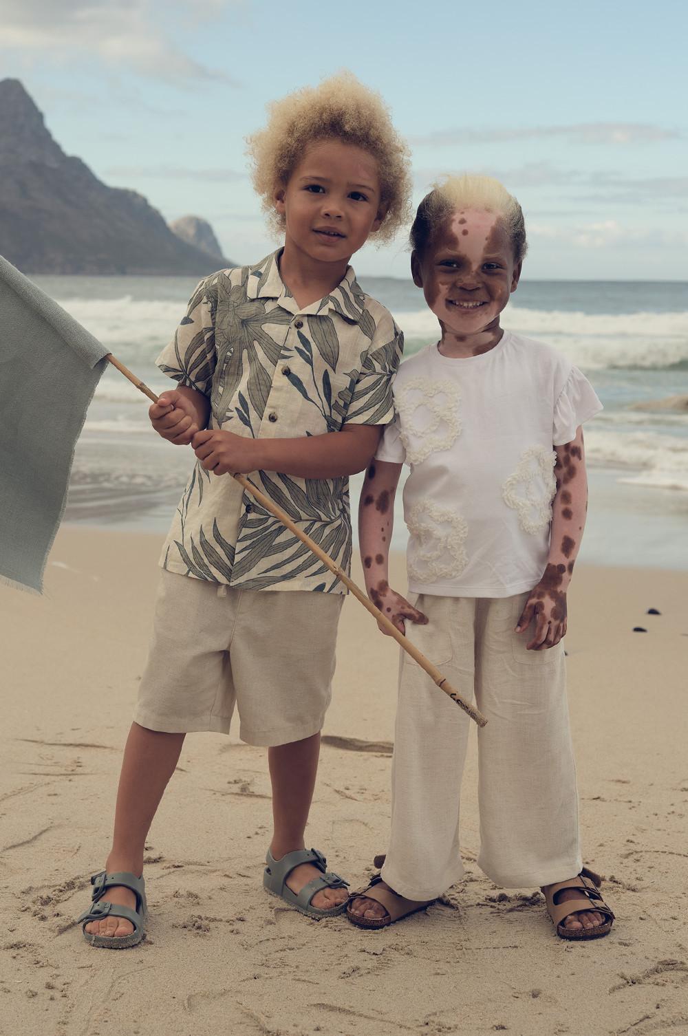 Zwei Kinder am Strand in heller Kleidung, eines in einem Hemd mit tropischem Print und Shorts, das andere in weißem Leinen.