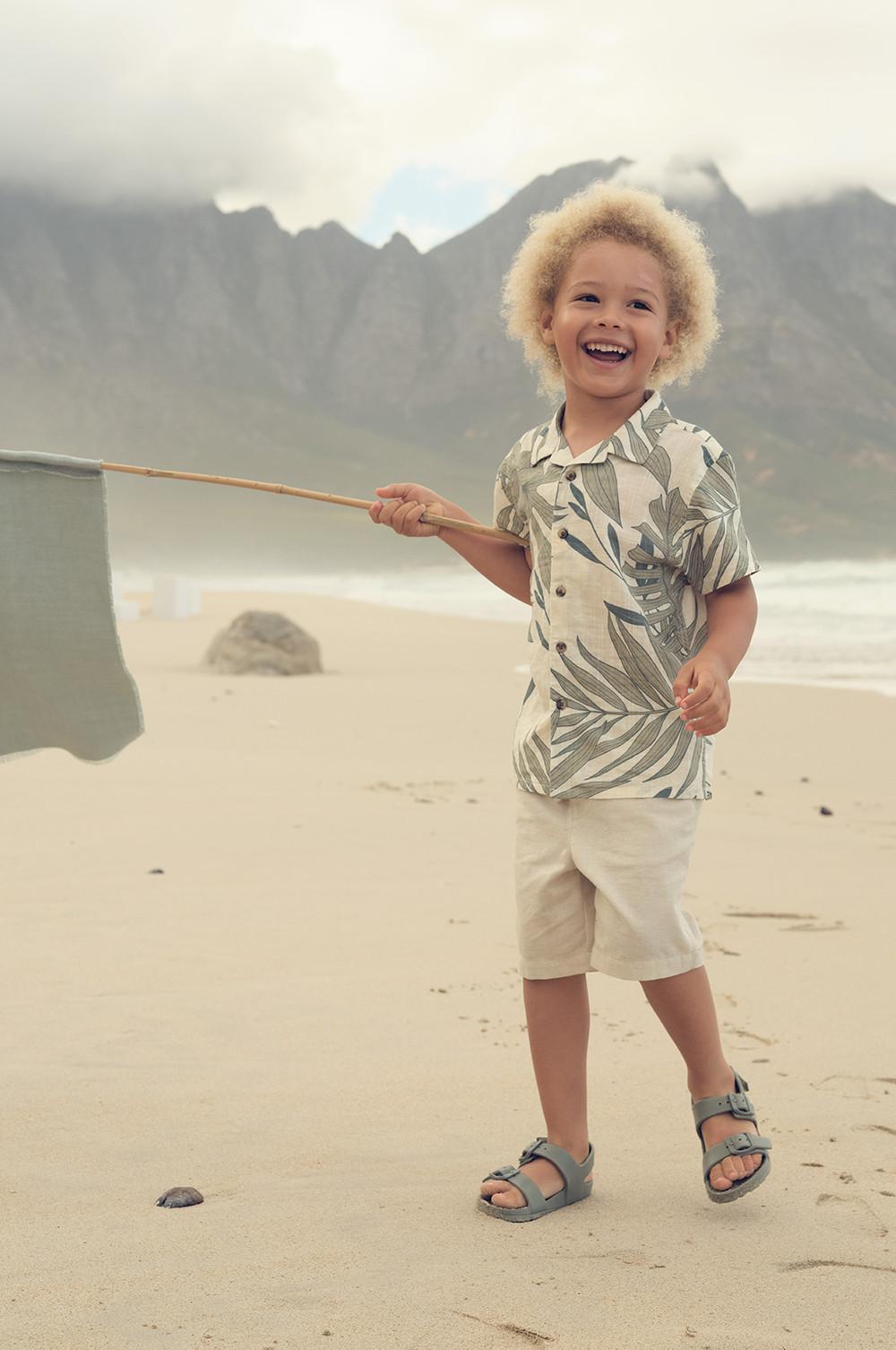 Kind in Hemd mit tropischem Print und khakifarbenen Shorts, das mit einer Flagge am Strand spielt, mit Bergen im Hintergrund.