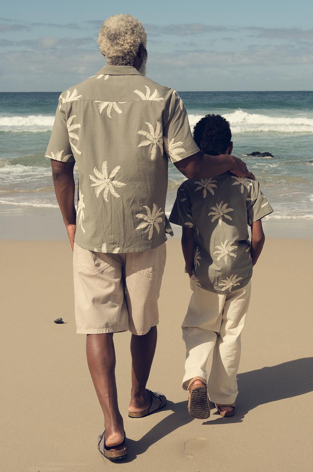Ein Erwachsener und ein Kind stehen am Strand, tragen zueinander passende Hemden mit Palmen-Print und helle Hosen und schauen zu den Meereswellen.
