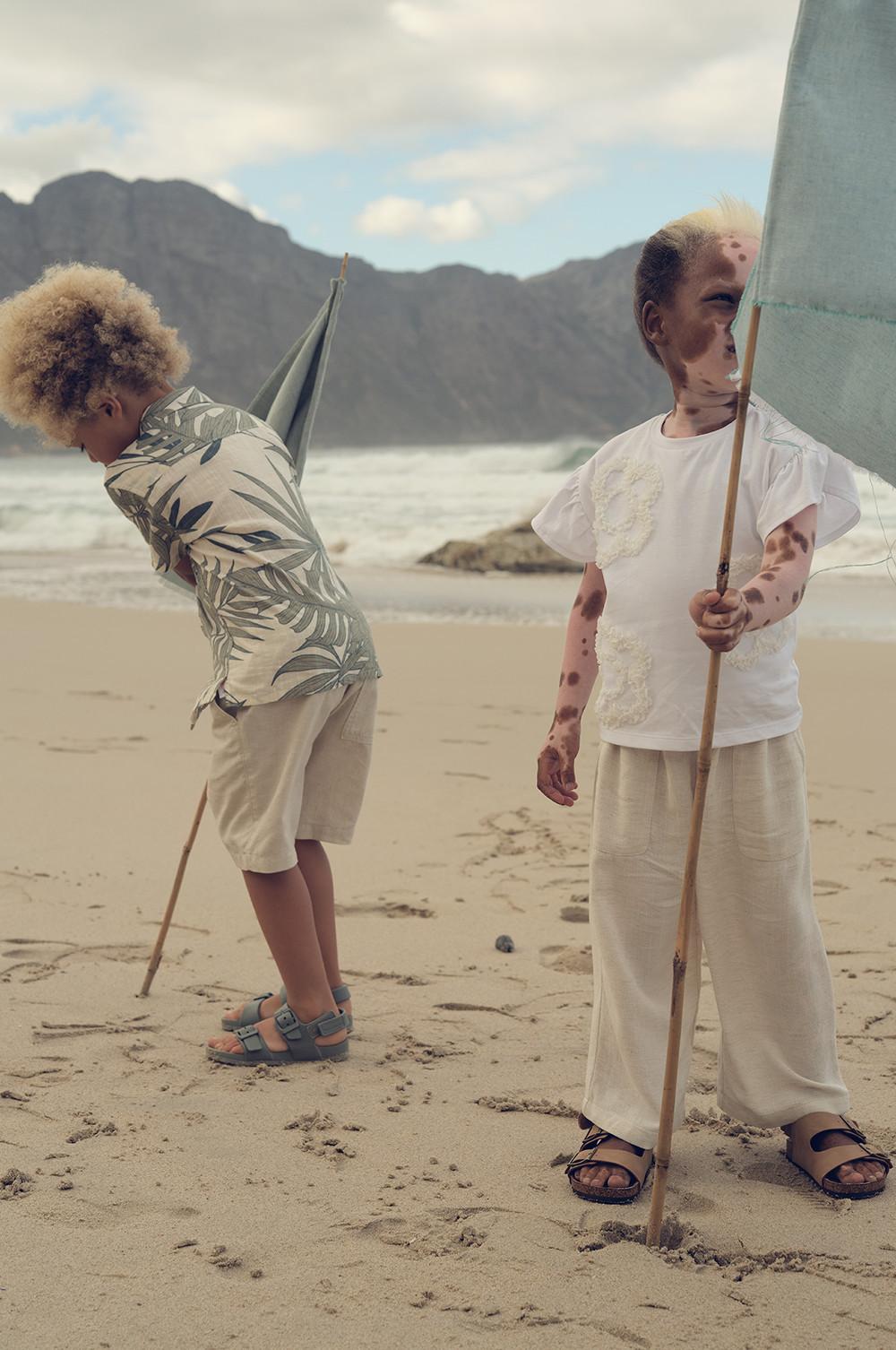 Zwei Kinder spielen am Strand mit Holzstöcken vor einer Bergkulisse, sie tragen lässige Sommerteile und Sandalen.