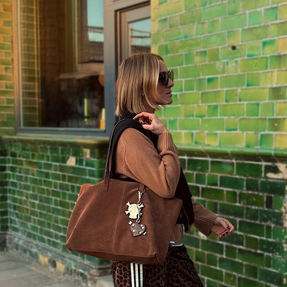 Model wearing brown sweater carrying a faux suede tote bag with decorative charms against vibrant green tiled wall.