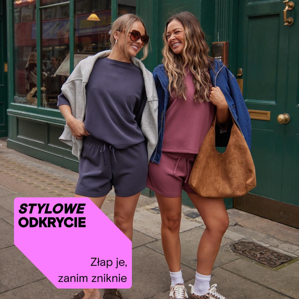 Two women smiling outdoors wearing casual matching top and shorts sets with hoodies and carrying bags.