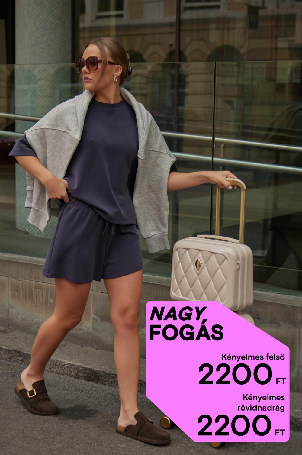 A napszemüveges modell sötétkék szabadidős felsőt és rövidnadrágot visel szürke kapucnis pulóverrel, és egy krémszínű bőröndöt húz.