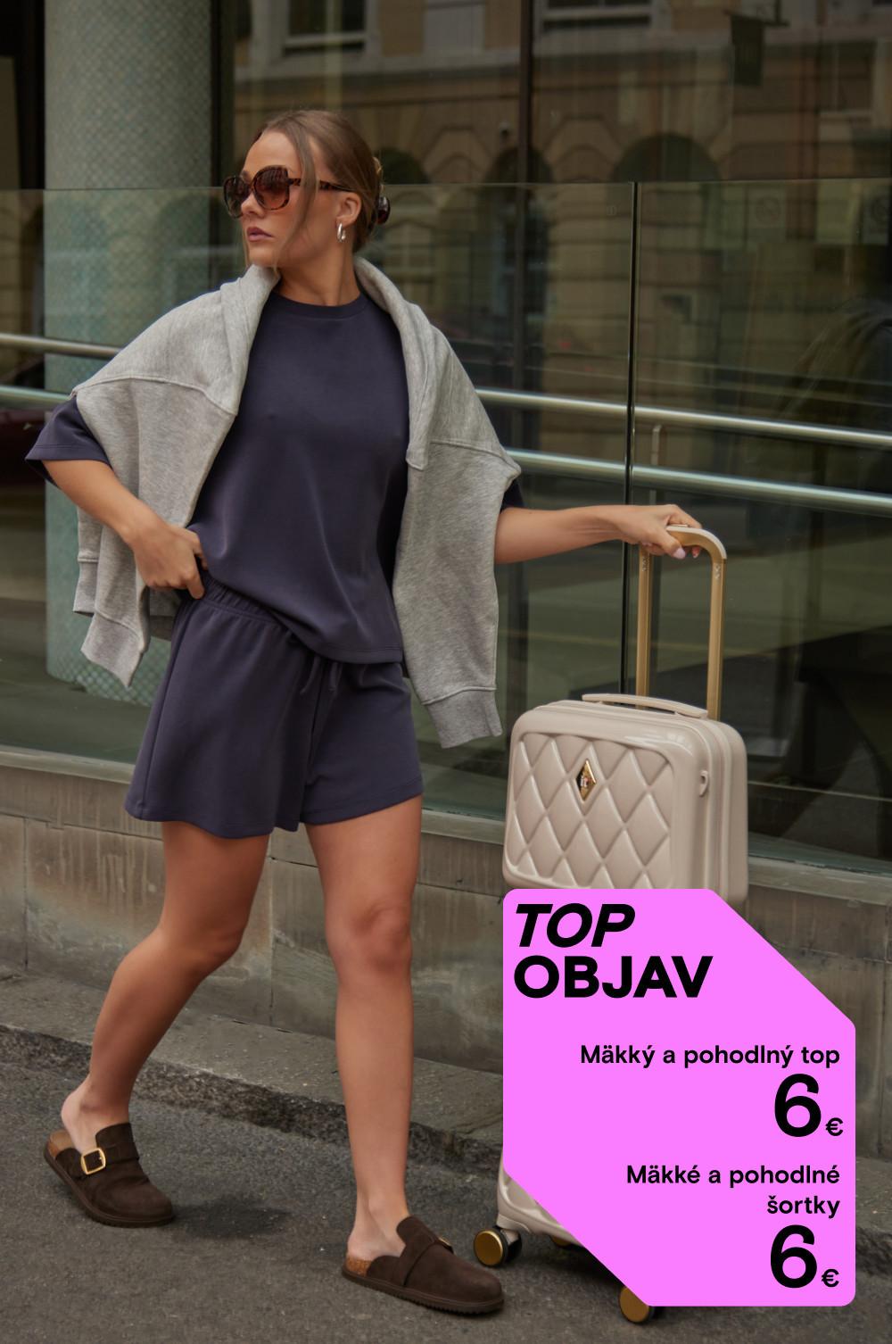 Modelka v slnečných okuliaroch má oblečený ležérny top so šortkami a sivou mikinou a ťahá za sebou béžový kufrík.