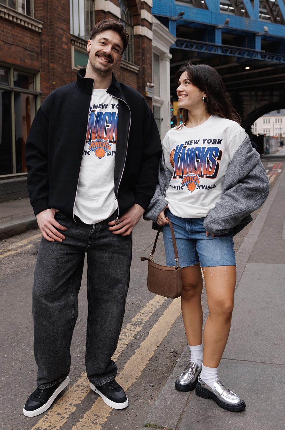 Two models wearing white New York Knicks graphic t-shirts, one styled with a grey bomber jacket, denim shorts and silver loafers. The other in black baggy jeans, a black bomber jacket and black trainers.