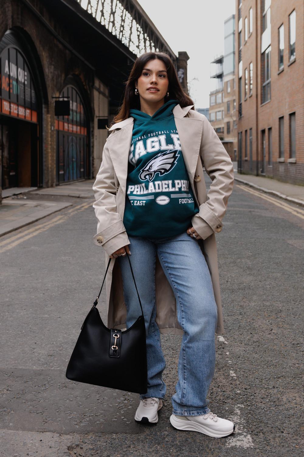 Model wearing Eagles Philadelphia hoodie, trench coat, jeans, holding black bag, walking on street.