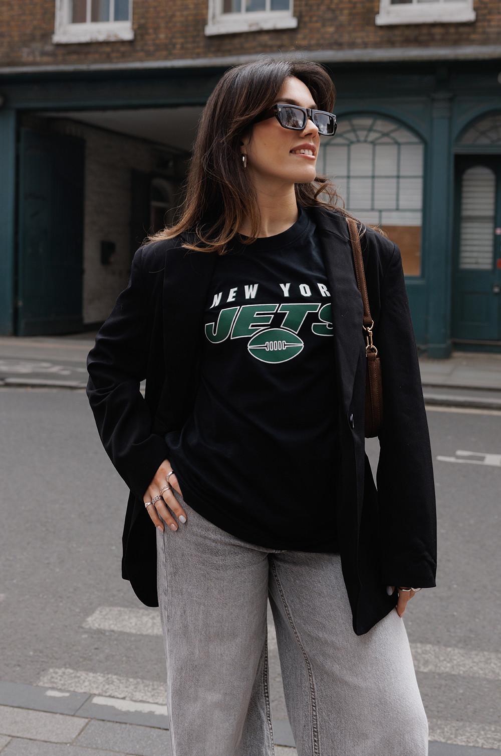 Model wearing New York Jets T-shirt, black blazer, grey wide leg jeans, holding a brown bag.