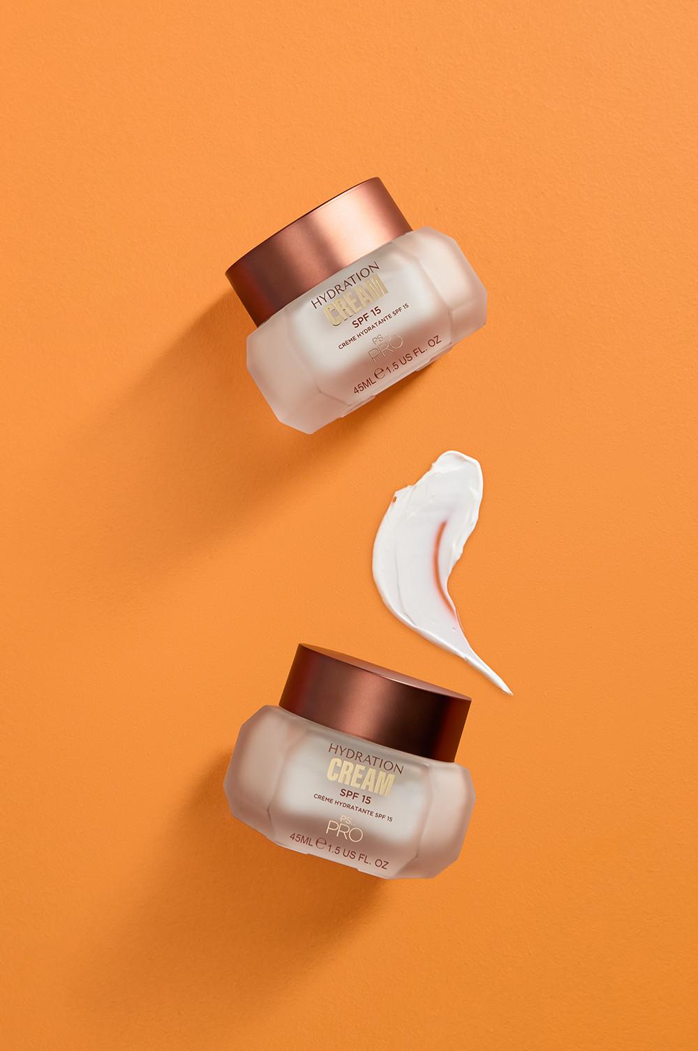 Due barattoli di crema idratante con SPF 15 su sfondo arancione, separati da una striscia di prodotto in evidenza al centro.