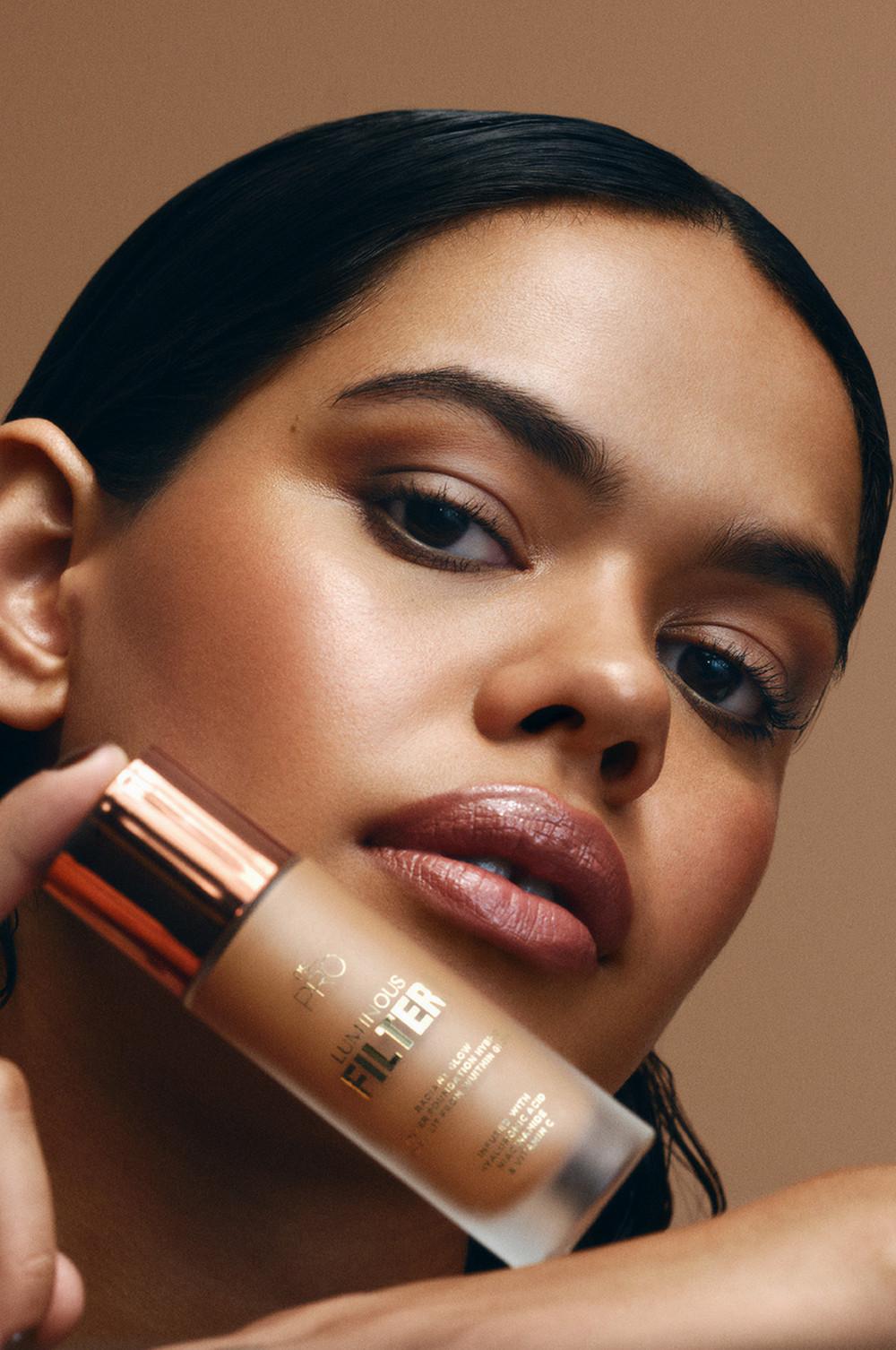 Modella che tiene in mano il fondotinta Filtro luminoso PS... Pro con applicatore, mettendo in evidenza il trucco liquido arricchito con acido ialuronico, niacinamide e vitamina C.
