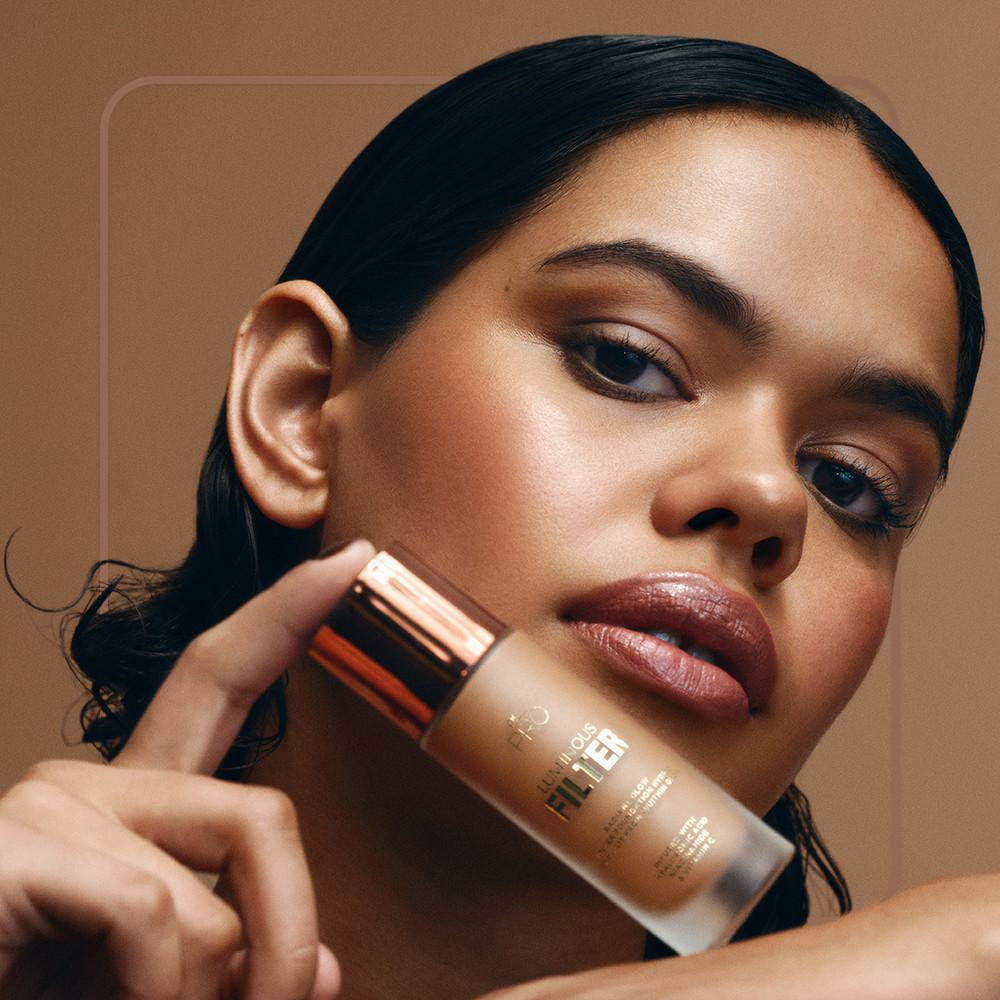 Modelo sujetando un frasco de base de maquillaje con tapa de color cobre junto a su rostro.