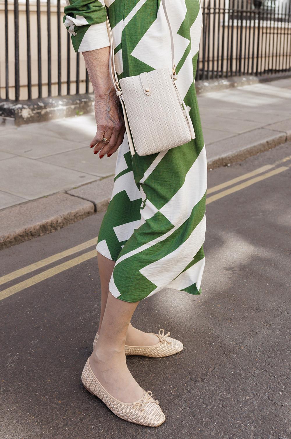 Model wearing green and white geometric print dress with cream woven crossbody bag and beige ballet flats standing on street.