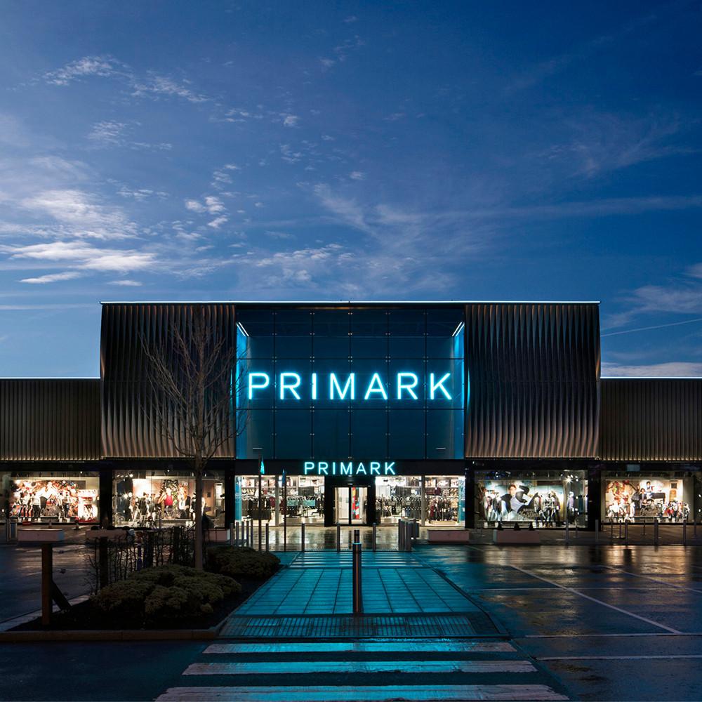 Primark store exterior illuminated at night with blue neon signage and display windows showing clothing collections.