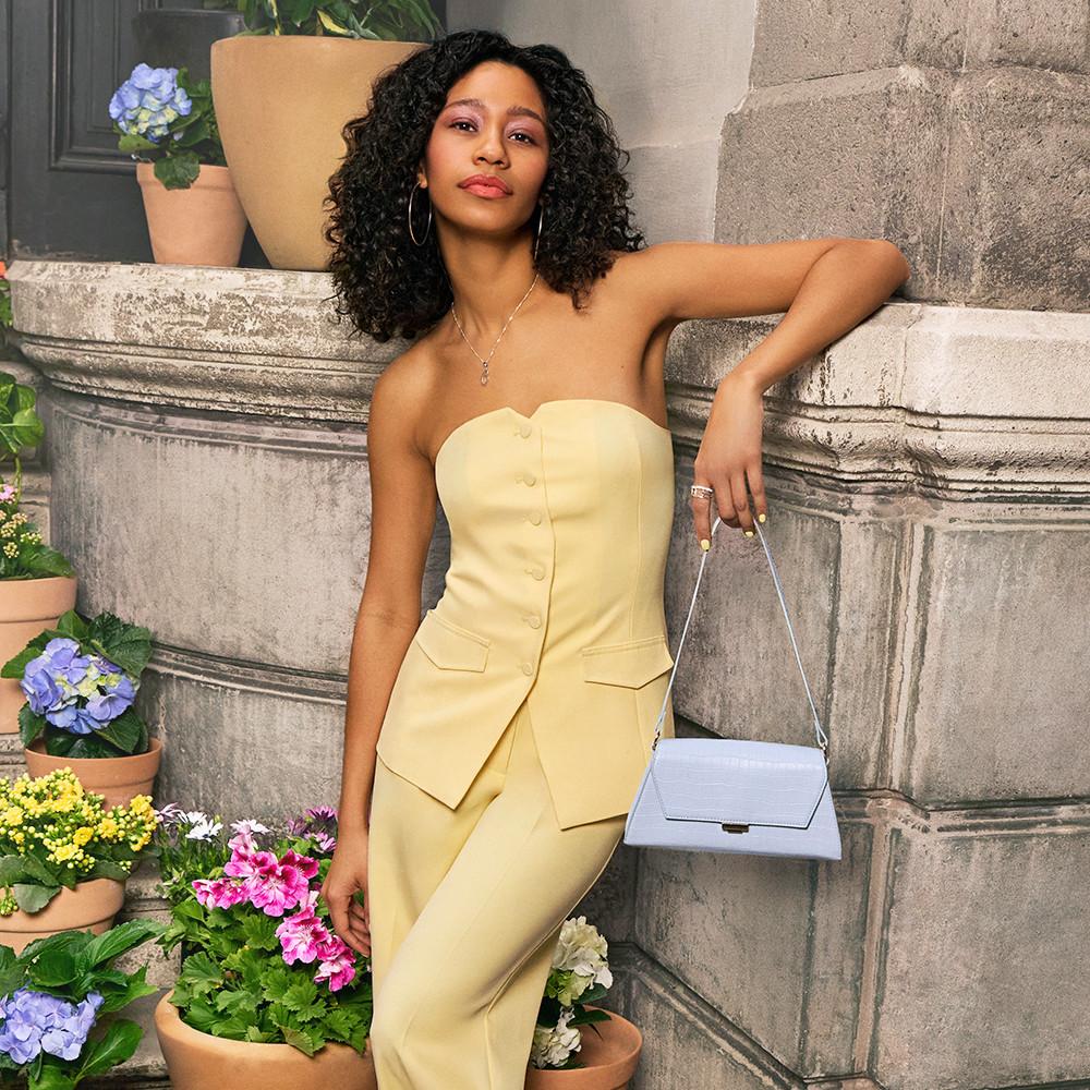 Model wearing butter yellow strapless waistcoat with button detail and pocket accents, paired with matching yellow tailored trousers, holding a light blue handbag against stone wall surrounded by potted flowers.