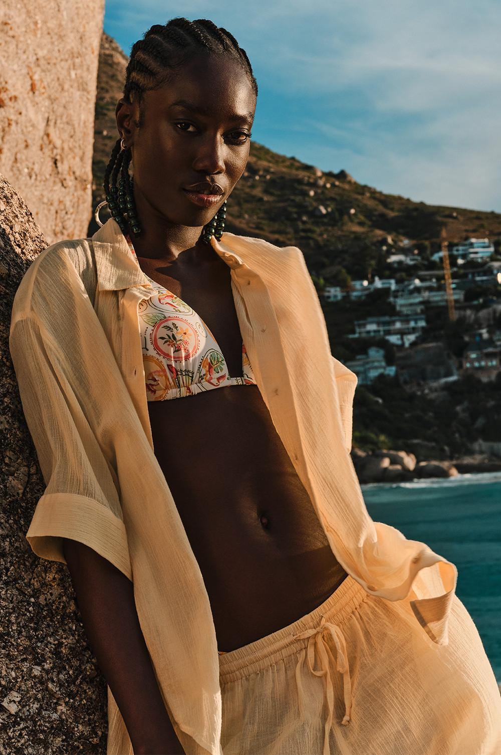Haut de bikini à motif avec une chemise de plage crème transparente et le bas assorti, sur fond de paysage côtier.