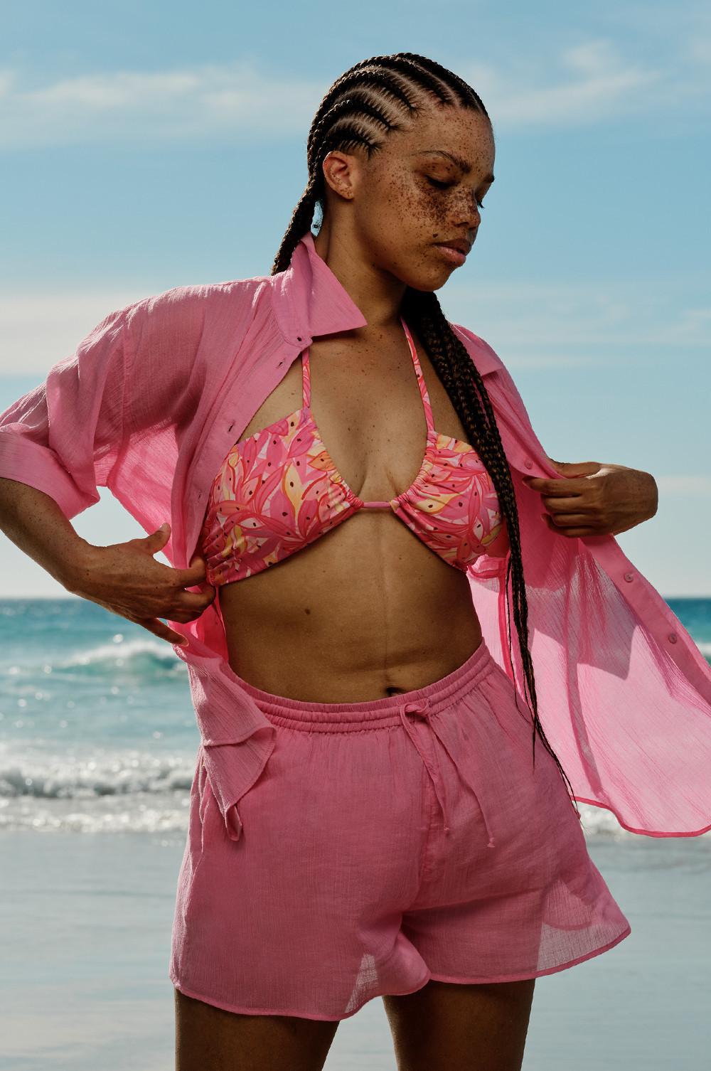 Tenue de plage rose avec haut de bikini imprimé, chemise boutonnée transparente et short assorti, l'océan en arrière-plan.