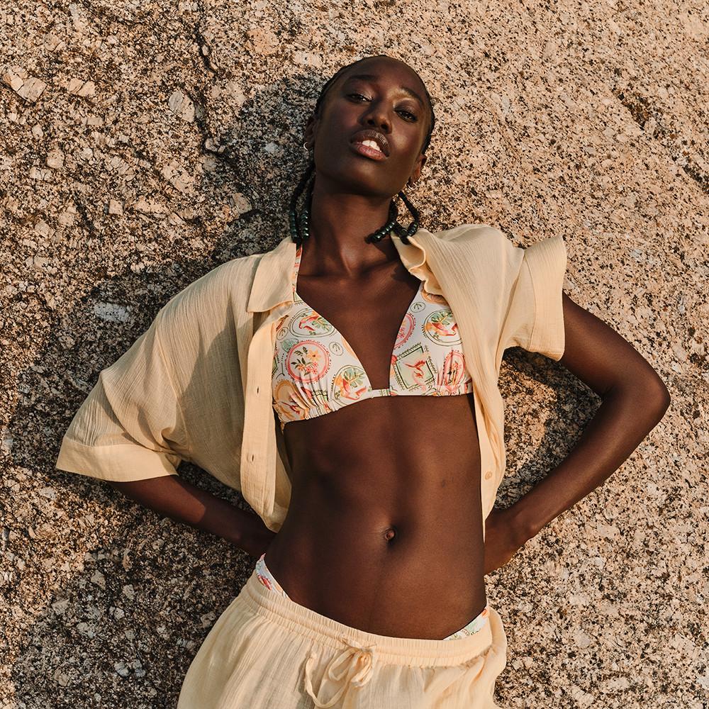 Haut de bikini triangle à motif associé à un short et à une chemise boutonnée crème légère, portée ouverte, le tout présenté devant une surface en pierre texturée.