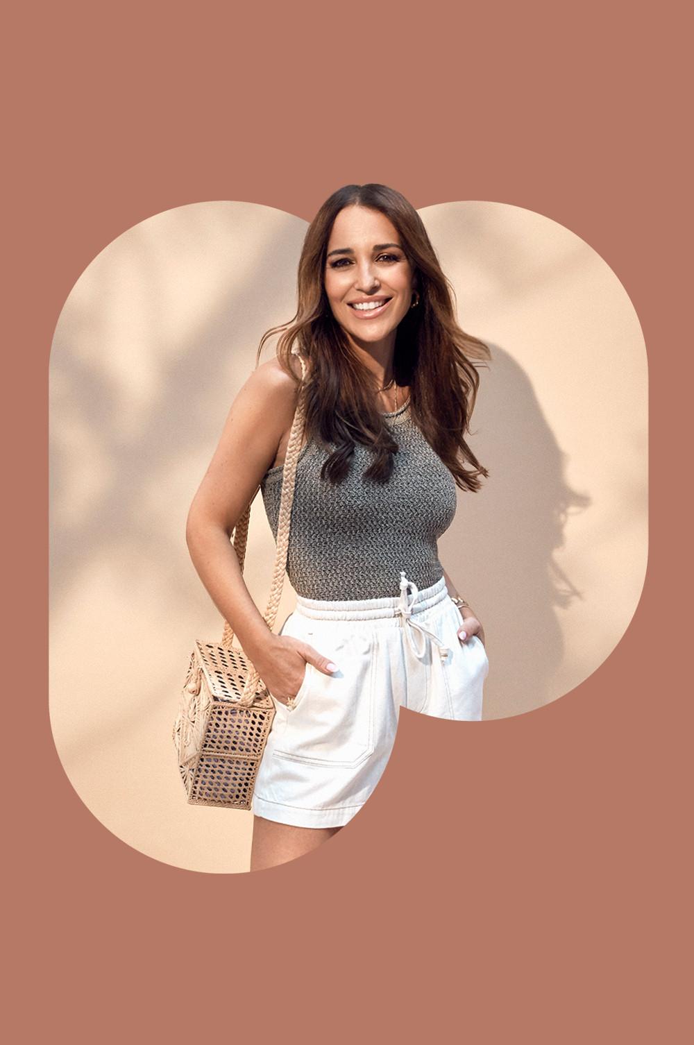 Paula Echevarria wearing gray knitted top with white drawstring shorts and woven straw bag against a terracotta background.
