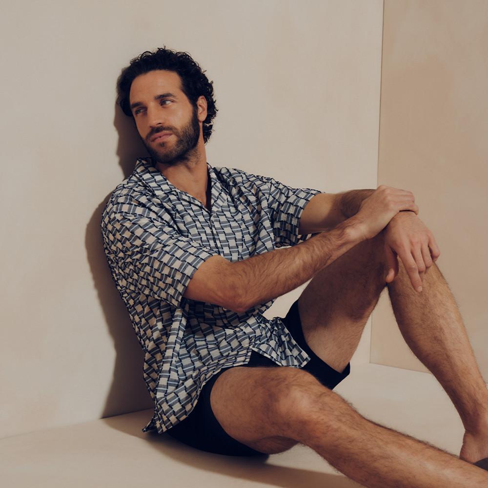 Model sitting wearing blue and beige geometric print short-sleeved shirt with black shorts.