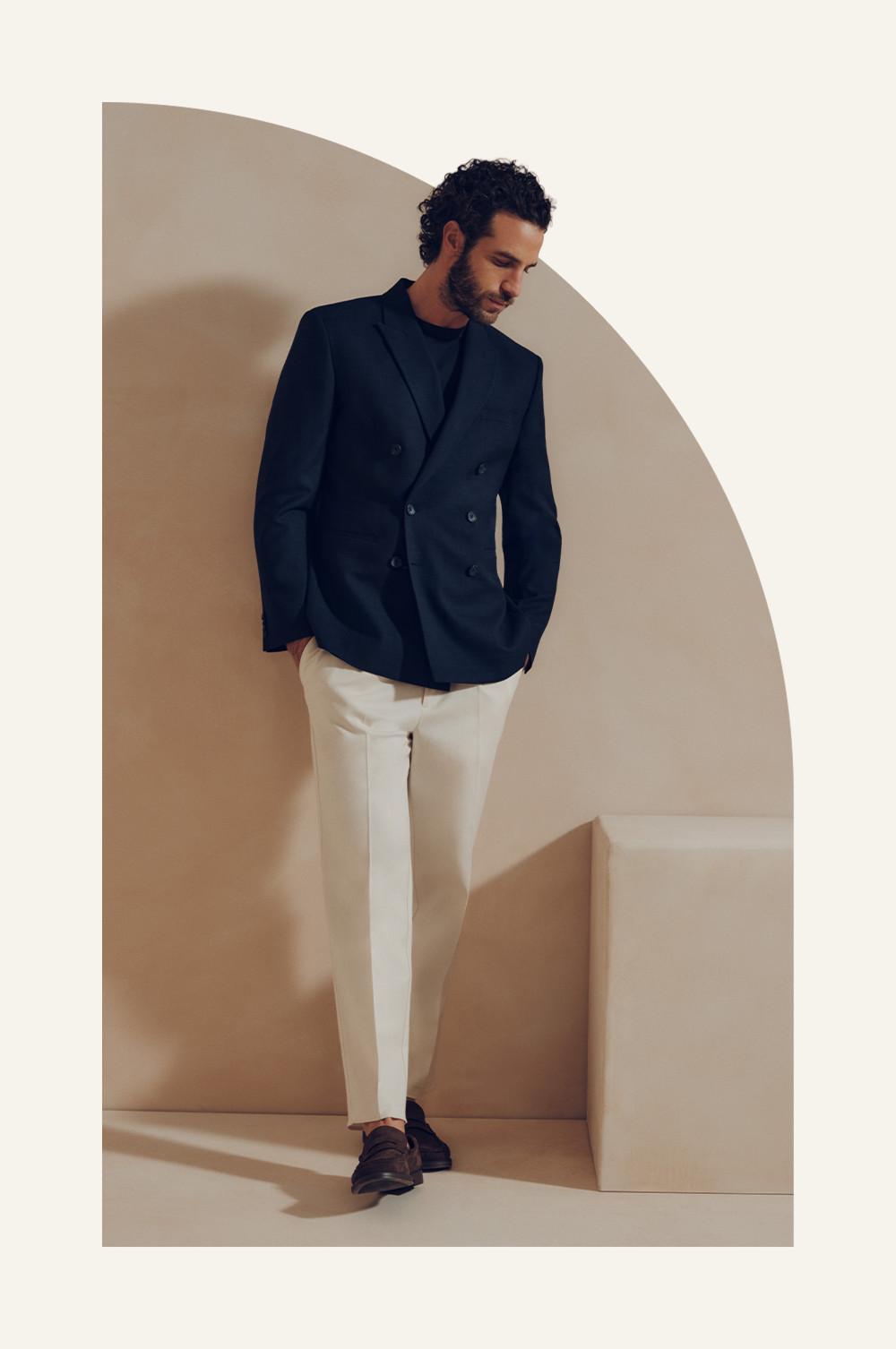 Model wearing navy double-breasted blazer with cream trousers and brown shoes, looking downward.