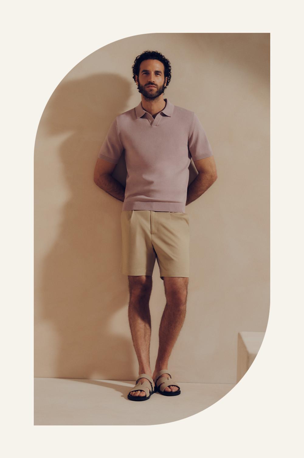 Model wearing mauve knitted polo shirt with beige shorts and sandals, standing in neutral archway.