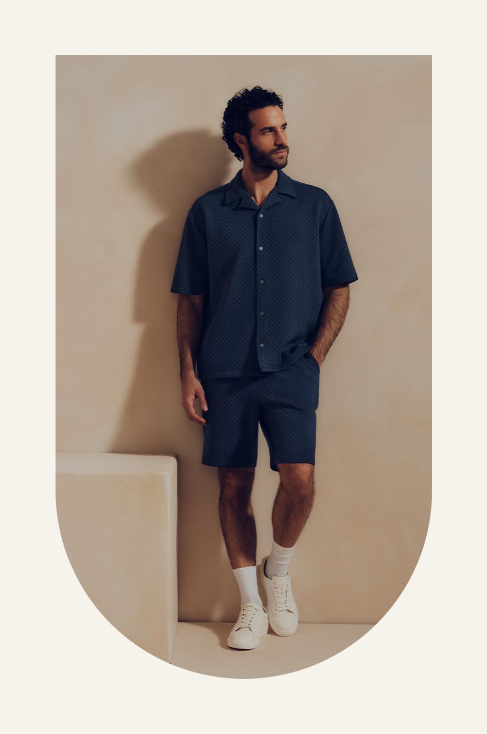 Model wearing navy textured short-sleeved shirt and matching shorts with white trainers and socks.