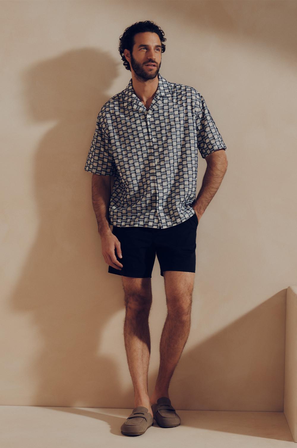Model wearing a geometric patterned short-sleeved shirt, navy shorts and loafers, standing casually.