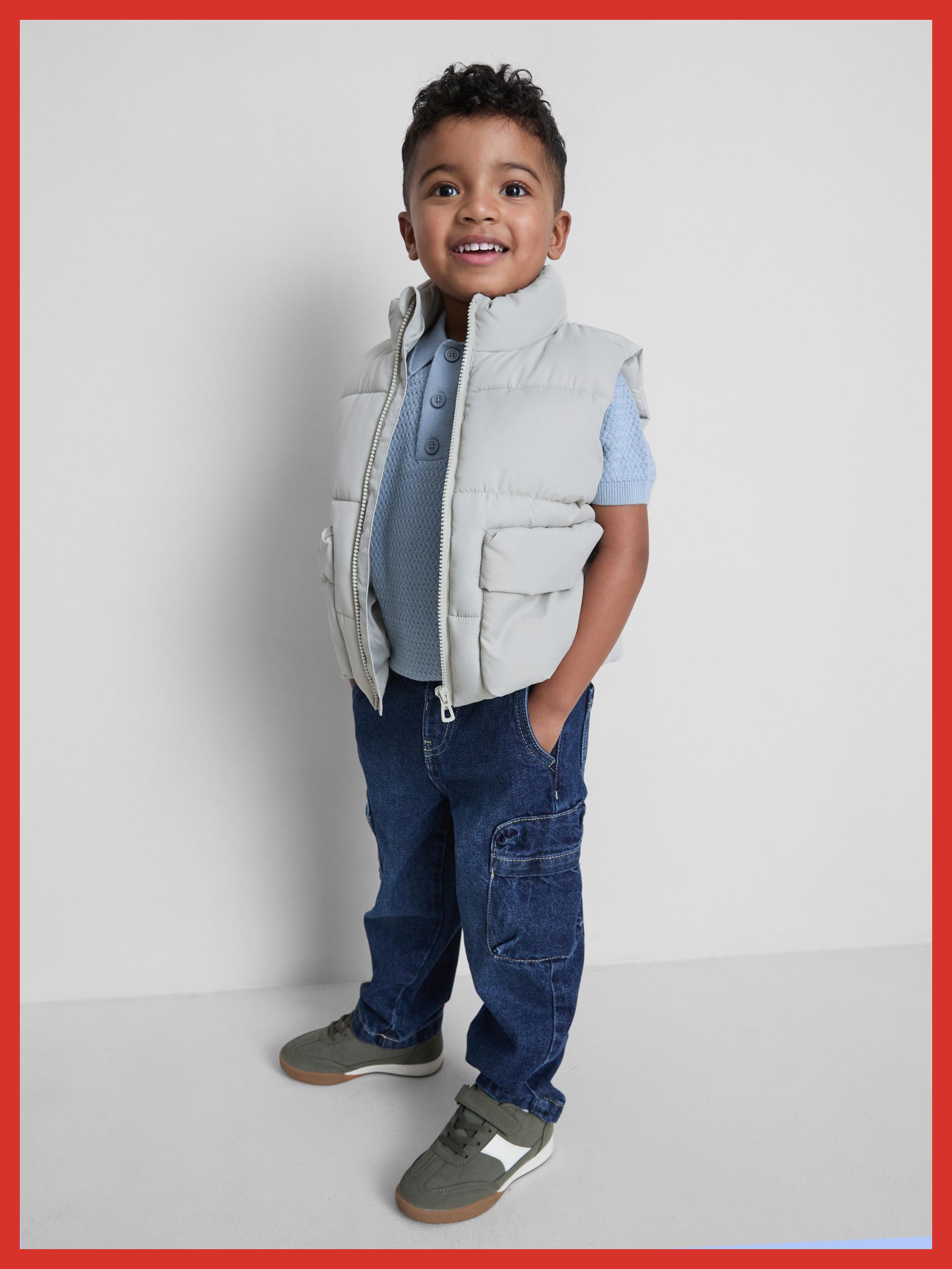Young boy smiling wearing a light grey padded gilet, blue polo shirt and dark blue cargo jeans.