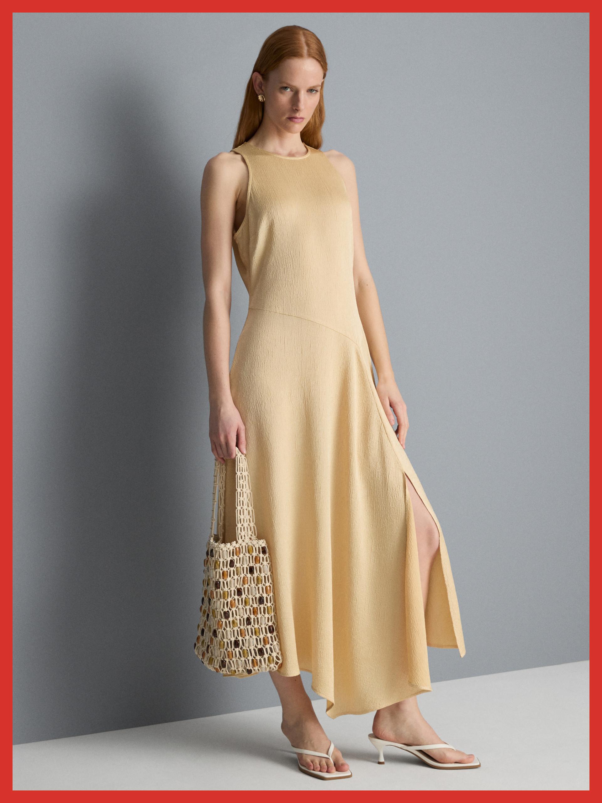 Model wearing a sleeveless butter yellow maxi dress with side slit, holding a woven tote bag.