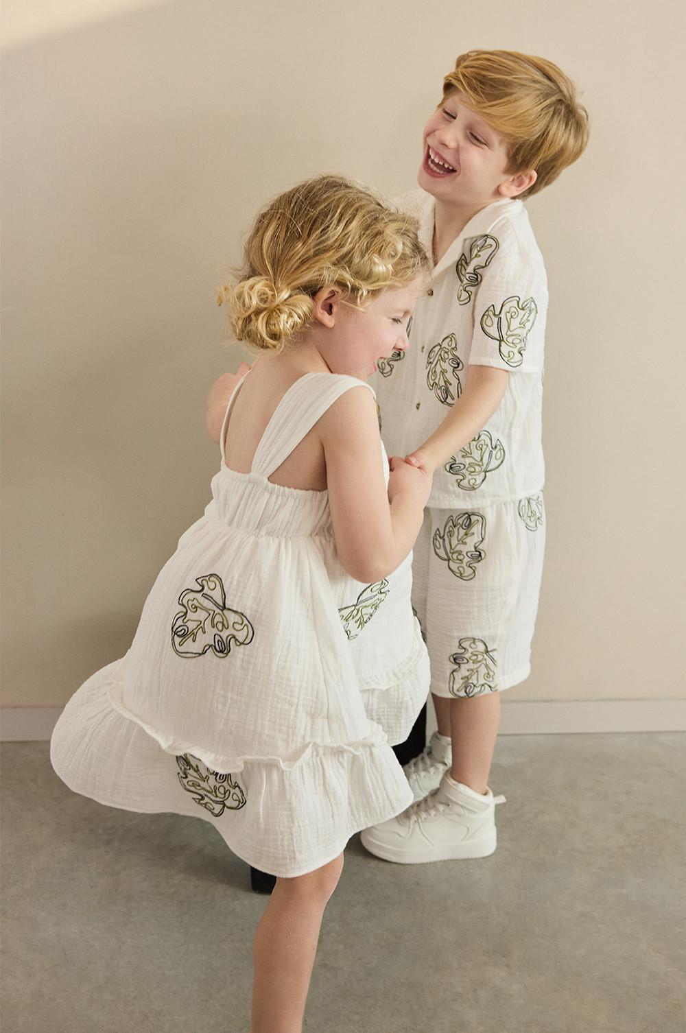 deux enfants, l'un portant une chemise blanche avec un short assorti, et l'autre une robe à motifs feuille sur l'ensemble