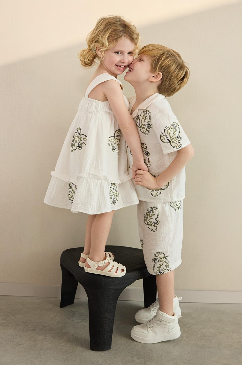 Dos niños, una con un vestido blanco bordado y sandalias y otro con un conjunto bordado de camisa y pantalones cortos y deportivas blancas.