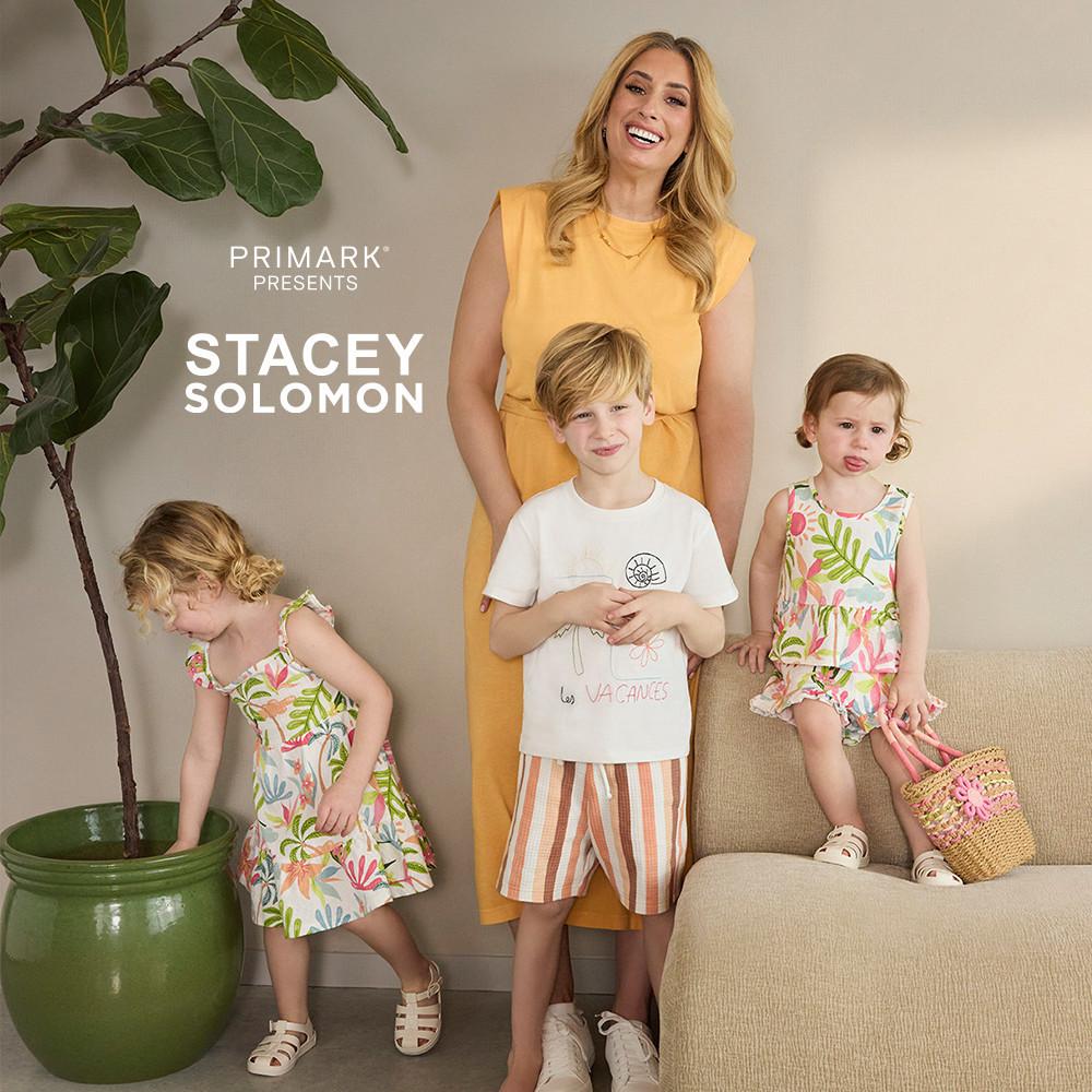Stacey Solomon con vestido amarillo junto a sus tres hijos, dos de ellos con conjunto estampado floral y vestido floral, y el otro con una camiseta blanca, pantalones cortos a rayas y zapatillas deportivas.