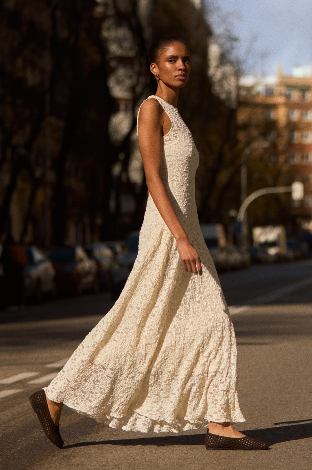 Model walking outdoors wearing a sleeveless cream lace maxi dress with flared skirt and woven flats.