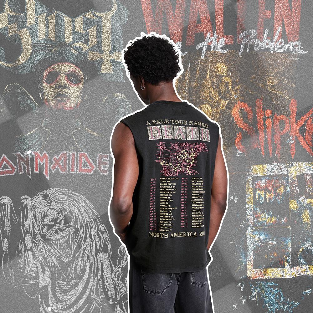 Person wearing black sleeveless tour shirt with tour dates printed on back, standing against graffiti wall.