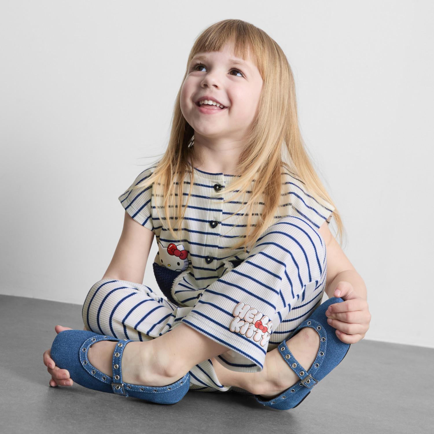 Young girl smiling wearing blue flutter sleeve top with heart pocket and matching striped trousers.