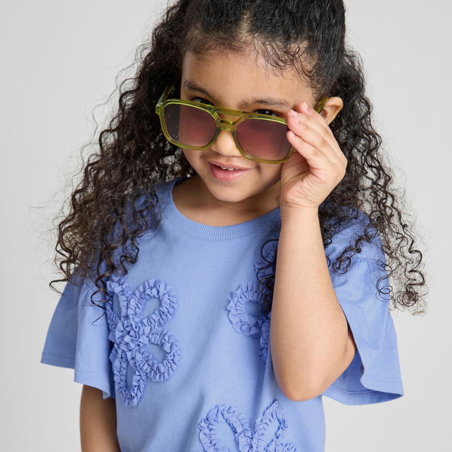 Girl with curly hair wearing oversized green sunglasses and blue ruffled top, smiling at camera.