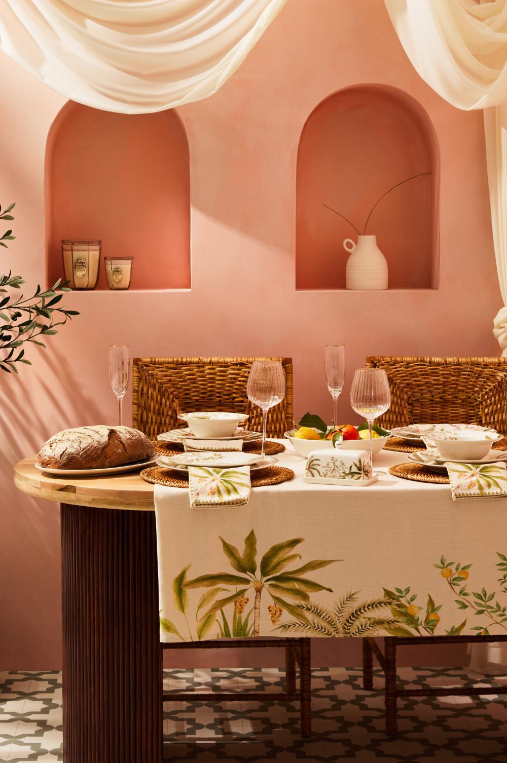 Dining table set with woven chairs, bread, plates, wine glasses and botanical print tablecloth in coral room.