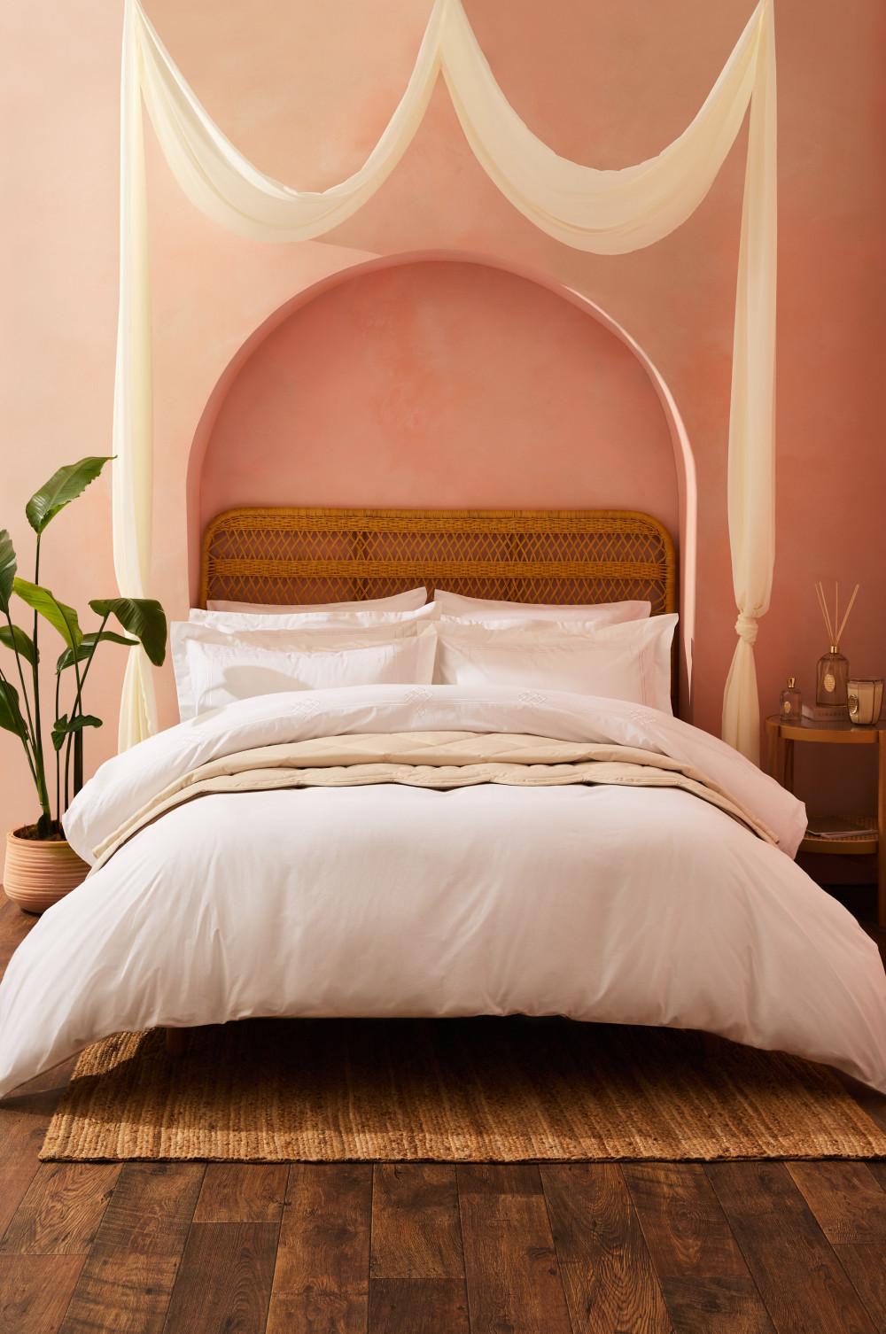 Bed with white and beige bedding, woven rattan headboard, set against a pink arched alcove wall.