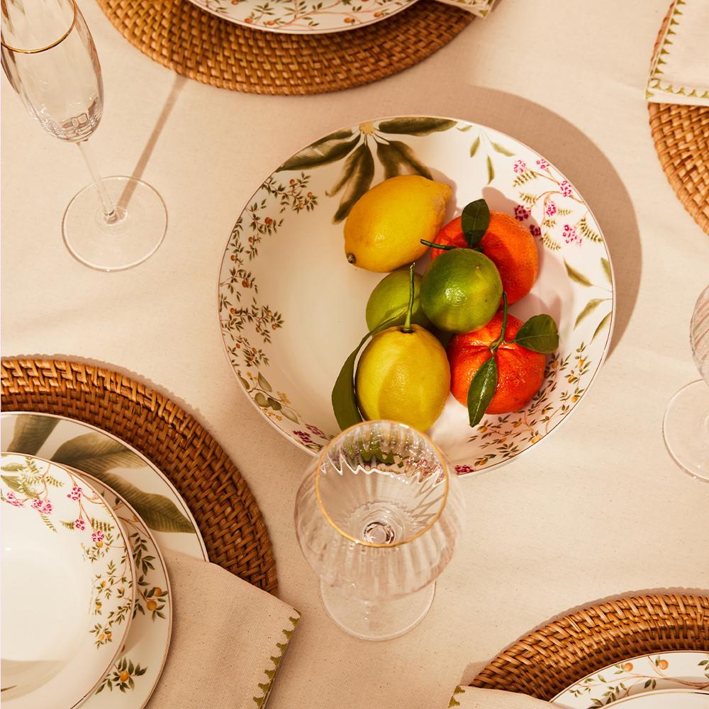 Decorative bowl with fresh citrus fruits displayed on a table setting with woven placemats and glassware.