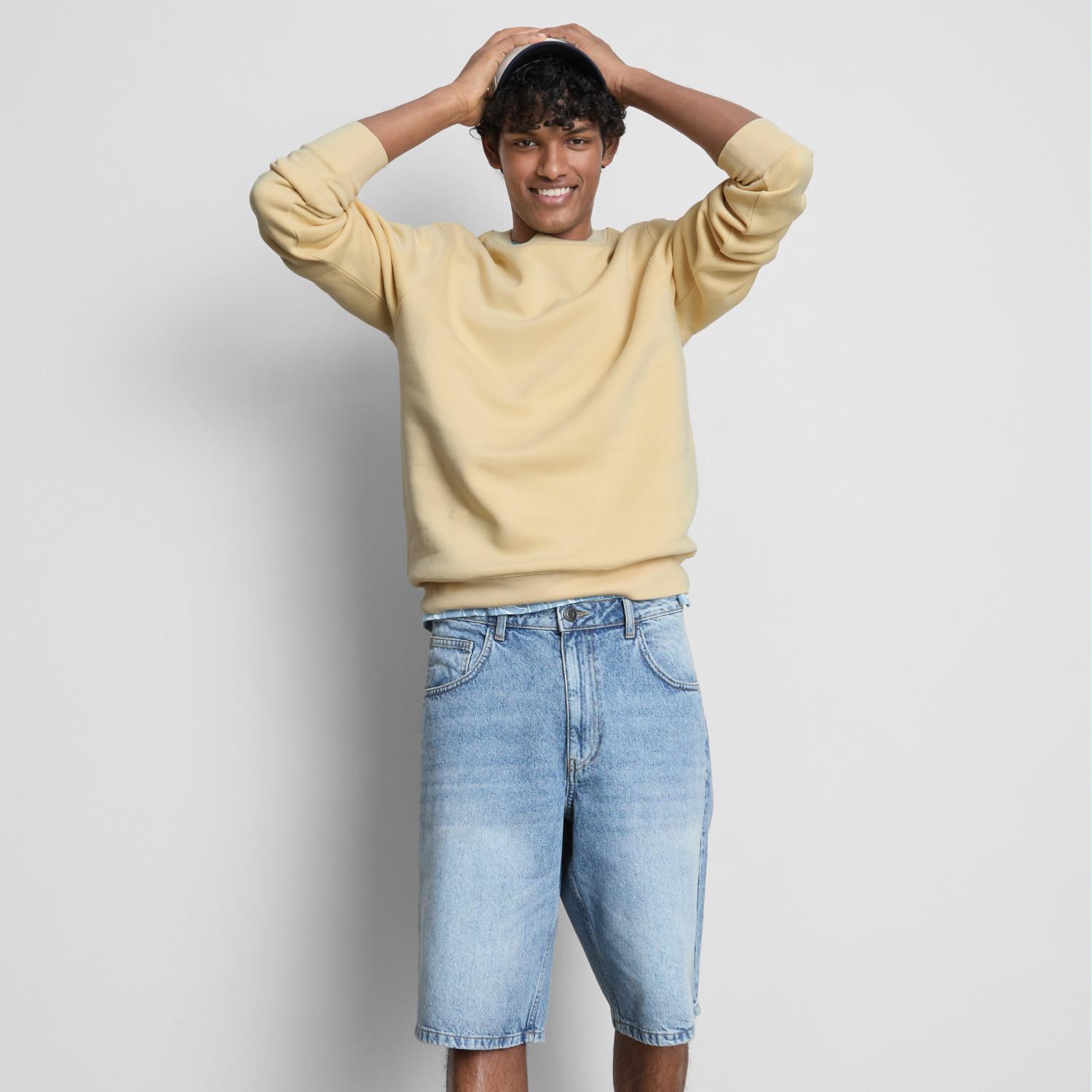 Model wearing pale yellow sweatshirt and light blue denim shorts with hands in hair, smiling.