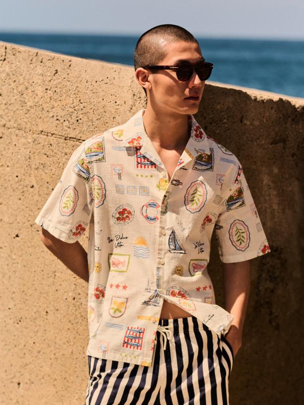 Man wearing sunglasses and cream short-sleeved shirt with travel-themed print, paired with striped shorts.